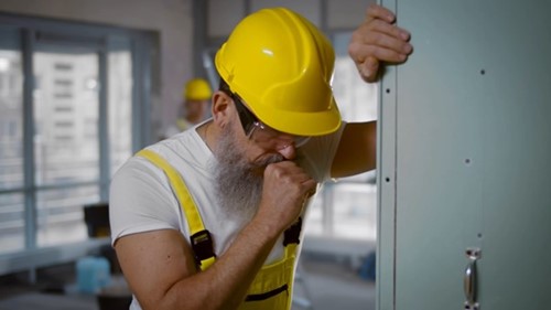 A construction worker coughing as a result of complications from workplace asbestos exposure and lack of personal protective equipment.