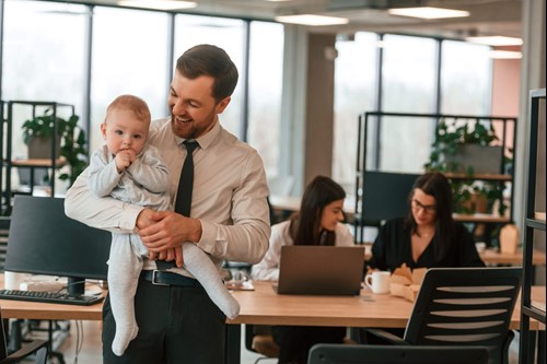 Working father holding a baby, whilst in an office surrounded by colleagues.